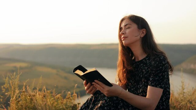 A long-haired girl reads a book in the sun. A young woman reads the Bible outdoors. A woman holds a Bible in her hands and studies the word of God on top of a mountain. 