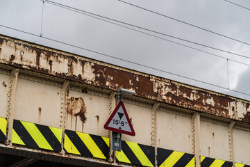 View of a weathered and rusted railway bridge with peeling white paint. The bridge features a triangular warning sign indicating a height restriction.
