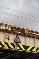 View of a weathered and rusted railway bridge with peeling white paint. The bridge features a triangular warning sign indicating a height restriction.
