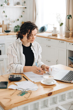Focused caucasian woman working at home in the kitchen with financial papers, counting on a calculator, paying bills, planning a budget to find a way to save money. Self-employed accounting