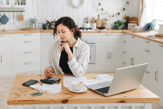 Focused caucasian woman working at home in the kitchen with financial papers, counting on a calculator, paying bills, planning a budget to find a way to save money. Self-employed accounting