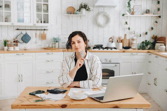 Portrait of young caucasian woman working at home in the kitchen with financial papers, counting on a calculator, paying bills, planning a budget to find a way to save money. Self-employed accounting