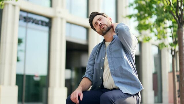 Mature man suffer from neck pain sitting on a bench near modern office building outdoors. Business male worker feels discomfort and back muscle Exhausted tired employee feels ache after sedentary work