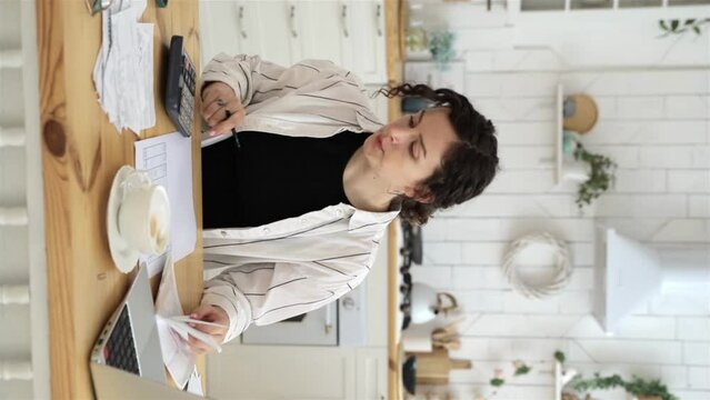 Vertical video. Caucasian woman working at home in the kitchen with financial papers, counting on a calculator, paying bills, planning a budget to find a way to save money. Self-employed accounting