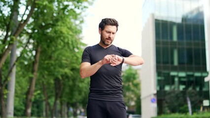 Sport man looking smart watch on the urban background of the city street. Man runner using smartwatch bracelet. Portrait fitness man checking result. Male in sportswear jogging satisfied with result