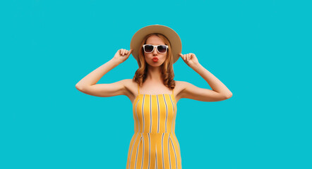 Summer portrait of beautiful young woman blowing her lips sending sweet air kiss wearing straw round hat on blue background