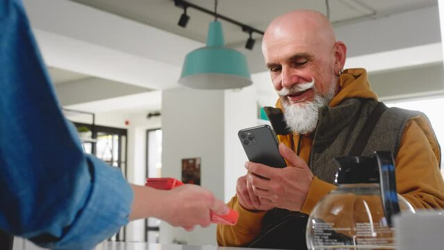 A modern old man with a beard and mustache pays with a smartphone with a qr code in a coffee shop or cafe. Senior buys coffee or tea take evey. Morning coffee for breakfast. Wireless technology
