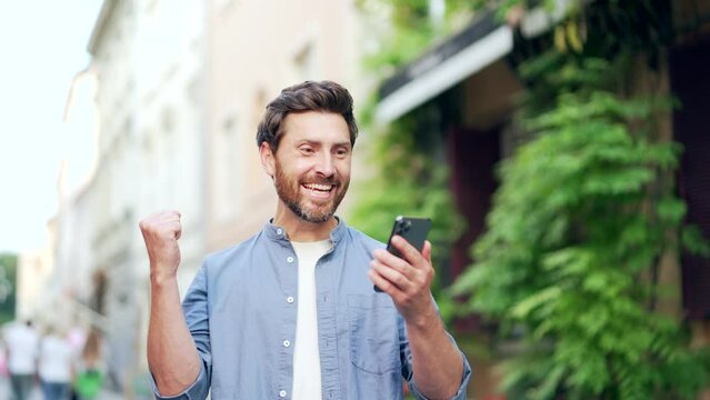Joyful mature adult man celebrating victory reading good great news on phone. Satisfied male receives a positive message looking at a smartphone while standing on a city street outdoors outside