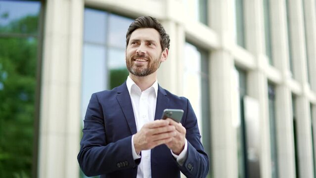 Happy businessman in suit uses mobile phone standing on city street outside near modern office building Bearded mature male entrepreneur manager or employee browsing chatting texting typing smartphone