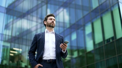 Confident mature bearded businessman in suit walking near modern glass business office building holding smartphone in hands. Man entrepreneur, investor, seo or manager using a mobile phone outside - Powered by Adobe