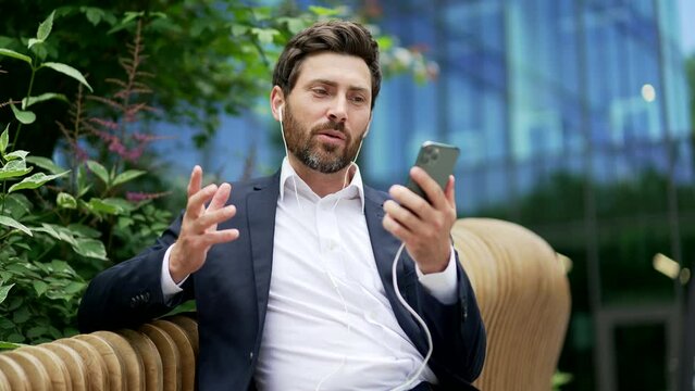 Cheerful businessman communicates online using mobile phone. Remote video call. Happy smile business man entrepreneur employee in suit talk sitting on bench during break near office building outside