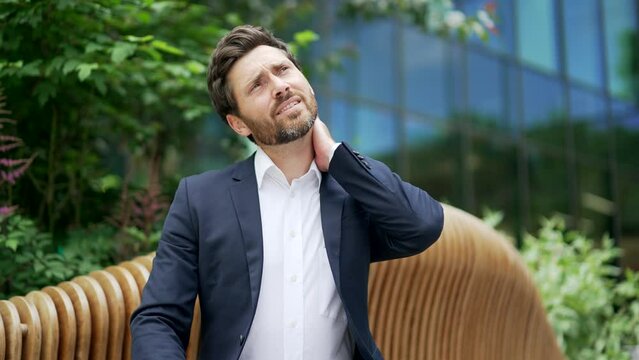 Employee suffer from neck pain sitting on a bench near a modern office building outdoors. Business male worker feels discomfort and back muscle pain Exhausted tired man feels ache after sedentary work