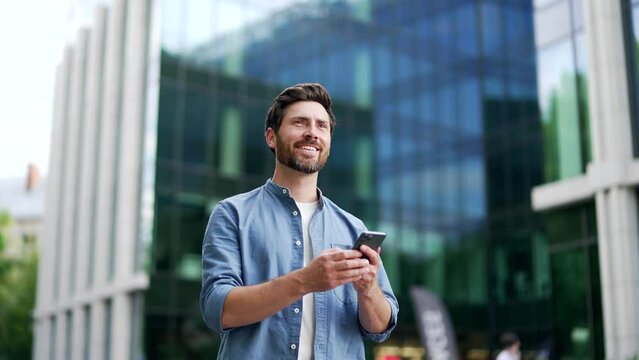 Handsome happy bearded businessman in casual walking near modern business office building hold smartphone in hands. Matureadult Man entrepreneur seo manager using read a mobile phone outside lifestyle