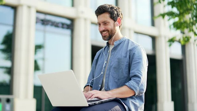 Handsome bearded businessman in casual working with laptop sitting on bench near modern office business center Happy Employee freelancer typing browsing laptop outside outdoors in city street smile