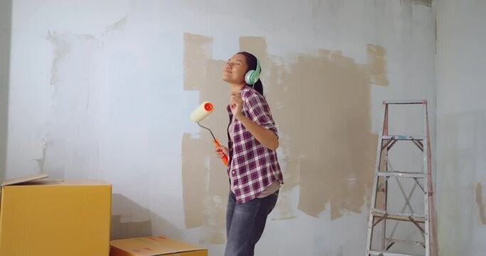 Young asian woman holding paint roller dancing at new home. Happy housewife making overhaul, home renovating, painting walls. Smiling young wife during major maintenance.