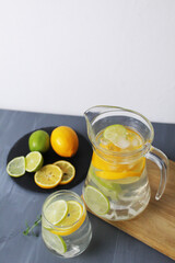 Citrus lemonade in a glass jug and a glass, lemon, orange, lime lie on a plate on a gray background. Summer refreshments, homemade lemonade
