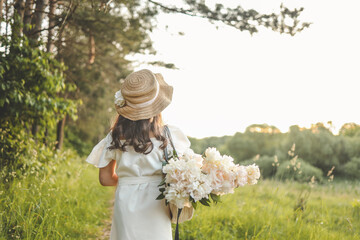 Little girl walking on the lawn with a bouquet of peonies and a hat