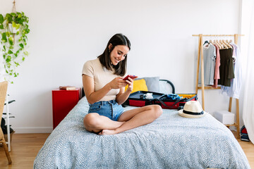Young pretty traveler woman using smartphone to book a hotel while preparing travel suitcase before...