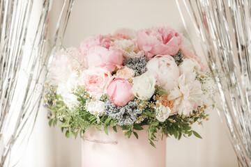 An elegant bouquet of fresh roses, peonies, and daisies in a pink box, beautifully arranged and placed by a light-filled window.