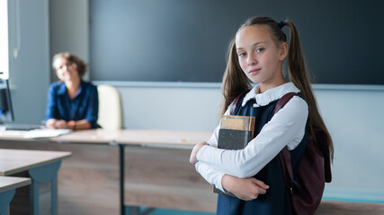 Fototapeta na wymiar Caucasian girl and female teacher in the classroom. The schoolgirl is holding a textbook.