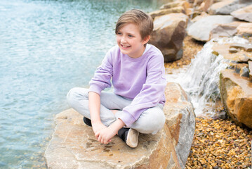 portrait of a happy child sits on huge stones near a mountain lake in front of a waterfall. joyful boy during summer holidays resting in the highlands