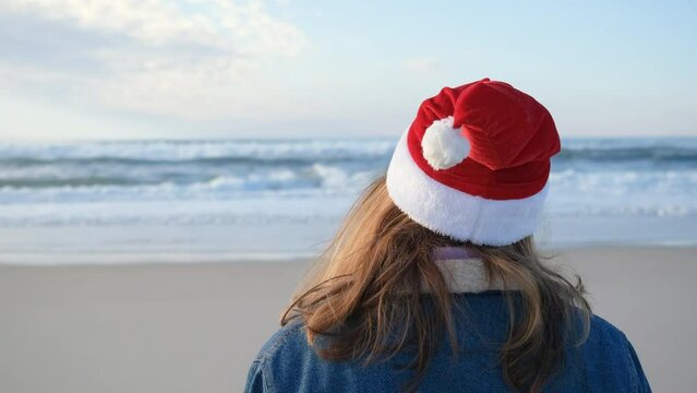 A girl or young woman in a red Santa Claus hat walks slowly along the shore of the Atlantic Ocean. Christmas or New Year holiday concept. 4K resolution video.