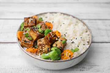 Bowl of rice with fried tofu, broccoli and carrots on white wooden table, closeup