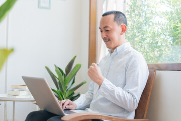 Young business man using laptop sitting at the chair in a home , looking at the paper, communicating online, writing emails, distantly working or studying on computer at home.