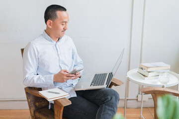 Young business man using laptop sitting at the chair in a home , looking at the paper, communicating online, writing emails, distantly working or studying on computer at home.