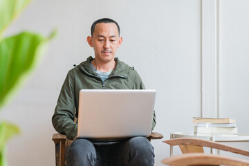 Young business man using laptop sitting at the chair in a home , looking at the paper, communicating online, writing emails, distantly working or studying on computer at home.