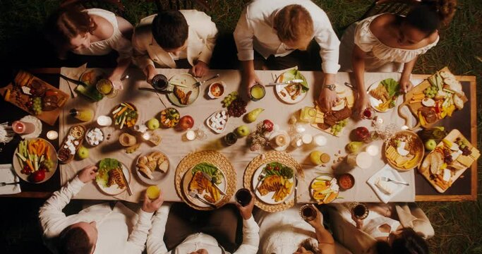 Guests clink glasses sitting at the beautifully served festive table. Top view, outdoor feast