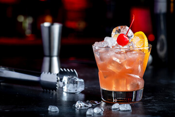 Matador alcoholic cocktail drink with scotch whiskey, white vermouth, liqueur, bitter, grenadine, orange juice, cocktail cherry and ice. Black bar counter background, steel bar tools and bottles