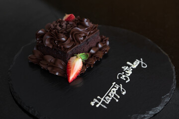 birthday dark chocolate cake or brownie cocoa cream and strawberries fruit with happy birthday text on black tray plate or dish for gift party greeting with sweet snack or dessert to bakery background