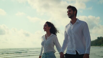 Young couple takes a leisurely stroll along the beach, their smiles beaming as they chat and laugh together, enjoying each other's company in the peaceful setting. Slow motion shot.