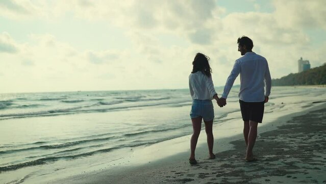 Young couple takes a leisurely stroll along the beach, their smiles beaming as they chat and laugh together, enjoying each other's company in the peaceful setting. Slow motion shot.