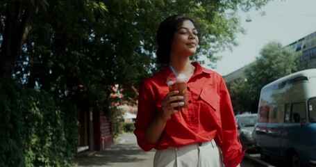 A young woman in casual clothes drinks lemonade from a disposable cup with a straw while walking around the city on a summer day