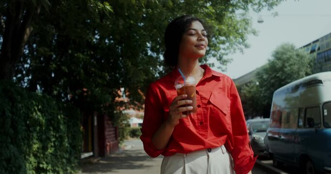 A young woman in casual clothes drinks lemonade from a disposable cup with a straw while walking around the city on a summer day