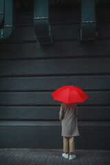 An unrecognizable woman under a red umbrella without a face stands with her back on the sidewalk....