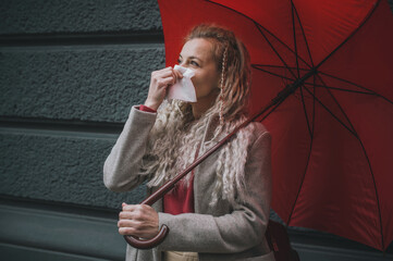 Mature blonde woman blows her nose in a napkin on a gray background under red umbrella on the...