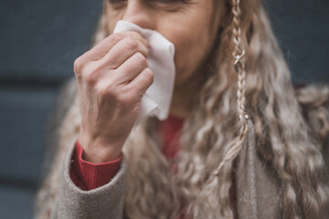 Mature blonde woman blows her nose in a napkin on a gray background. Autumn cold and seasonal drop...