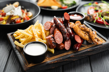 Grilled sausages served on cutting board with french fries and sauces on dark wooden background, top view.