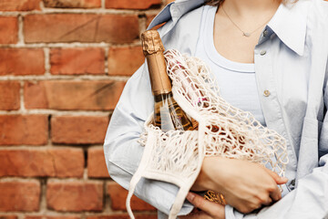 Young woman holding champagne sparkling wine in mesh bag in hands, she wearing in shirt and jeans,...