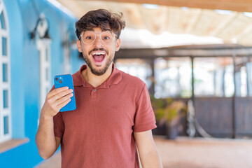 Young Arabian handsome man using mobile phone at outdoors with surprise and shocked facial expression