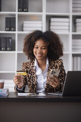 Young African American businesswoman holding credit cards with smartphone sitting at a desk in office. shopping online,