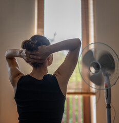 A young woman with her hands behind her head in front of a fan escaping from the summer heat. Back...