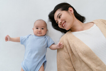 Top View of happy smiling Asian mother and newborn baby lying in bed with love in bedroom. Cute baby looking at camera near her mom at home. Enjoy tender family moment, motherhood, childcare concept.