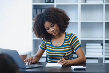 Young African American woman using laptop and Taking notes on a notebook sitting at desk in office.