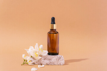 Brown cosmetic serum bottle on stone podium and white jasmine flowers on neutral beige background. Closeup