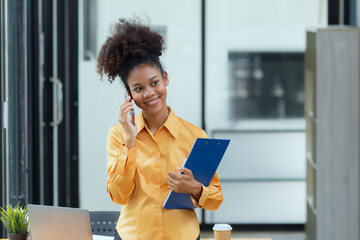 Young African American businesswoman using smartphone with clipboard standing in office. African American businesswoman.