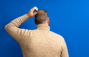 Back view of thoughtful man scratching his head over blue background. He wonders why he doesn't...
